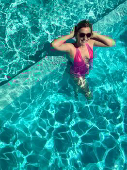 Young beautiful woman swims in the pool. Happy smiling girl in a pink swimsuit and sunglasses put her hands behind her head and looks at the camera. photo