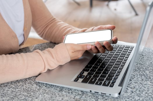 A woman in a cardigan using her smartphone while working on her laptop at a table indoors. photo