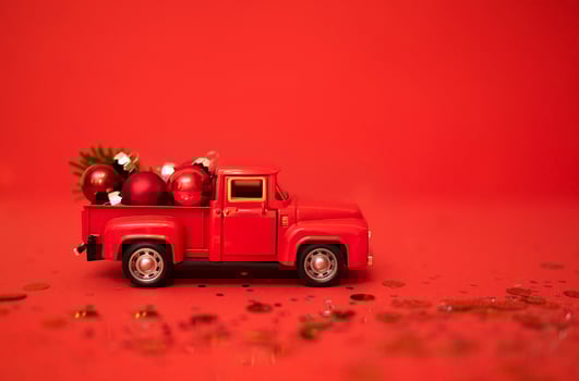 Red toy truck with Christmas balls and fir branch in its trunk. Miniature car with Christmas decoration on red background. Bright backgdrop with sparkles and confetti. photo