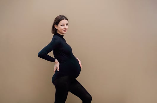 Young pregnant woman standing against beige background looking to the camera touching her tummy photo