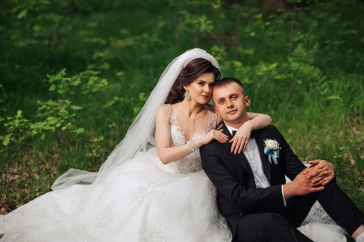 A bride and groom are sitting on the grass, with the bride wearing a white dress and the groom wearing a black suit photo