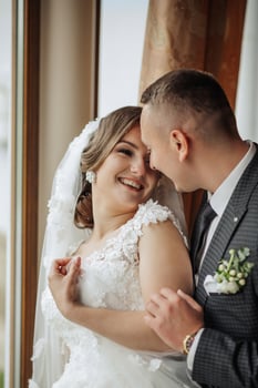 A bride and groom are hugging and smiling at the camera. The bride is wearing a white dress and the groom is wearing a suit. Scene is happy and joyful, as the couple is celebrating their wedding day photo