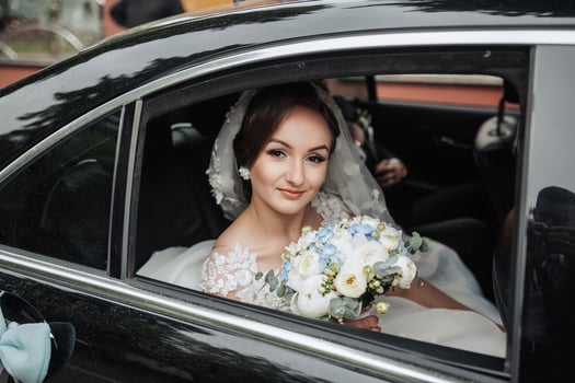 A bride is sitting in a car with a bouquet of flowers. She is smiling and looking out the window photo