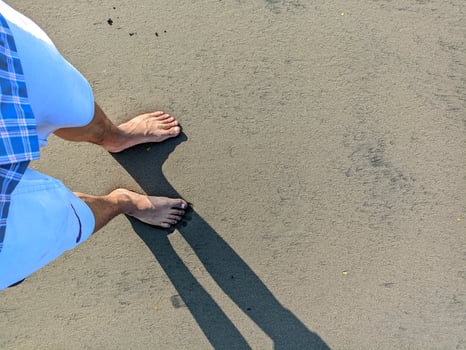 photo of a man's feet stepping on gray beach sand with his shadow
