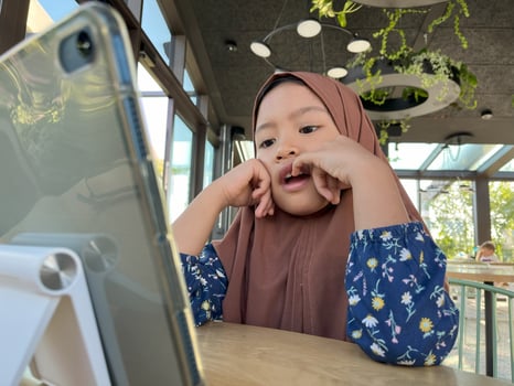 Happy cheerful Asian little Muslim girl siting and learning online with mobile tablet in cafe or restaurant. photo