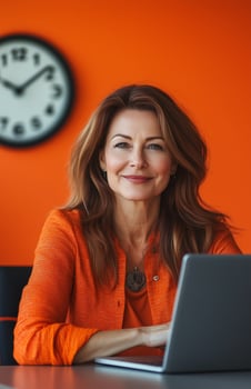 A smiling middle aged woman in an orange office, seated at her desk with a laptop photo