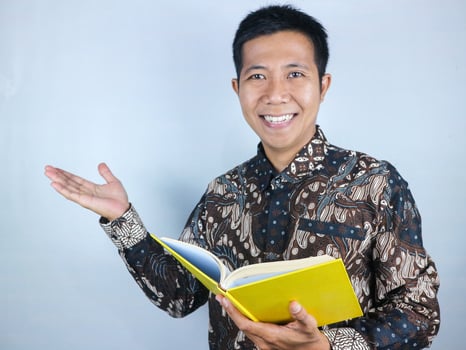 Smiling asian man wearing batik clothes presenting or showing something while holding a book photo