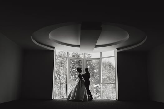 A couple is standing in front of a window, with the man wearing a suit and the woman wearing a wedding dress. The image has a mood of elegance and romance photo
