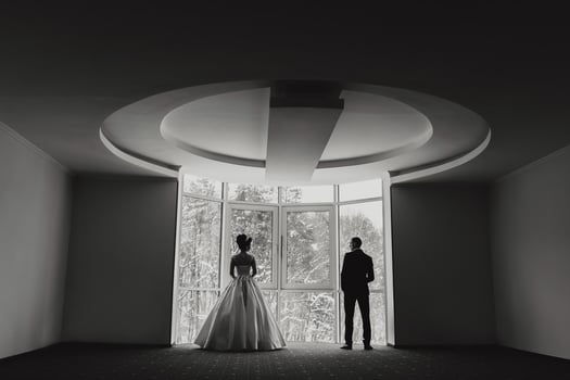 A man and a woman are standing in a room with a large window. The woman is wearing a white dress and the man is wearing a suit. Scene is one of anticipation and excitement photo