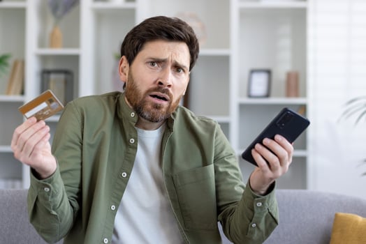 Confused adult man holding smartphone and bank card in hands while shrugging with worried expression. Uncertain customer having struggles with using ecommerce banking app for making online payments. photo