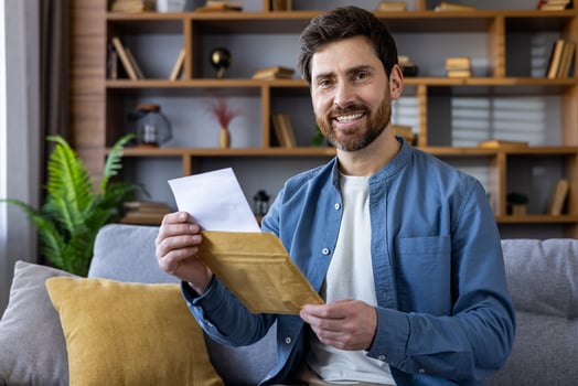 A young adult male in a casual outfit sits on a sofa, attentively opening a yellow envelope with a white letter inside. photo