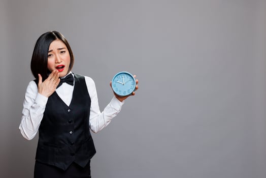Tired asian woman wearing waitress uniform yawning and covering mouth while holding alarm clock portrait. Sleepy receptionist running late for work, showing time and looking at camera photo
