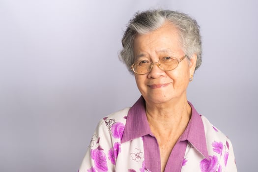 Beautiful elderly Asian woman with short gray hair wearing glasses and looking at the camera with a smile while standing on a gray background. Space for text. Aged people and health care concept photo