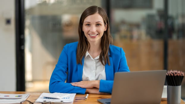 A young, cheerful professional woman sitting at her desk in a bright office environment, looking confidently at the camera. photo