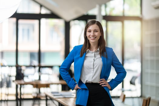 A professional woman with a confident smile stands in a bright, airy office space, dressed in a smart blue blazer with arms crossed. photo