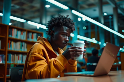 In Library, Young African American Man Works on Laptop, Sips Coffee photo