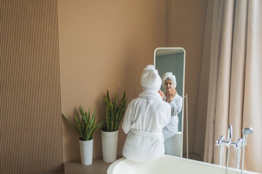 Mature adult old woman looking at mirror applying nourishing anti age face cream doing daily beauty routine. Senior woman with white mask creme on skin gets facial treatment Skin care aging skincare photo