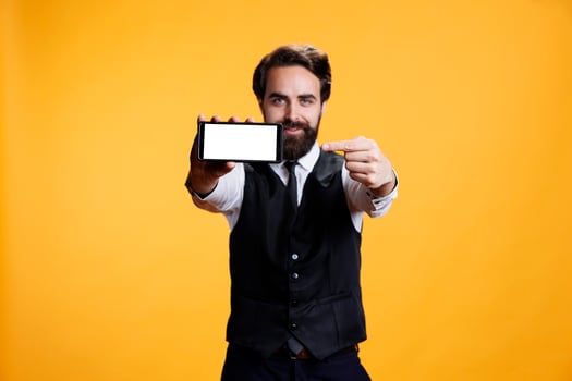 Skilled butler presents white blank display against yellow background, showing isolated mockup empty screen. Male waiter using smartphone with copyspace template in studio. photo