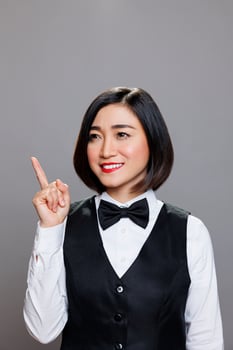 Cheerful asian woman wearing waitress uniform directing upwards with finger and posing in studio with joyful facial expression. Restaurant smiling receptionist pointing up photo