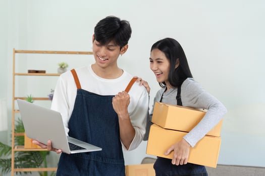 Young Couple Selling Products Online from Home, Smiling and Excited, Holding Laptop and Cardboard Boxes photo