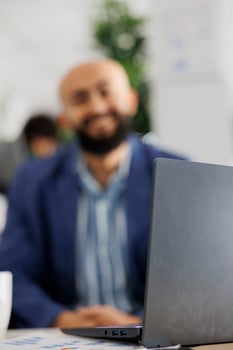 Blurred company employee working on laptop in business office. Smiling arab man start up entrepreneur using portable computer at workplace in coworking space and looking at camera photo