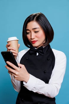 Hotel asian woman receptionist enjoying break while chatting on smartphone and drinking takeout coffee. Young waitress in uniform holding tea paper cup and texting on mobile phone photo
