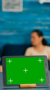 Isolated digital tablet with mock up green screen chroma key display standing on office desk in living room. In background room collegues talking about online communication photo
