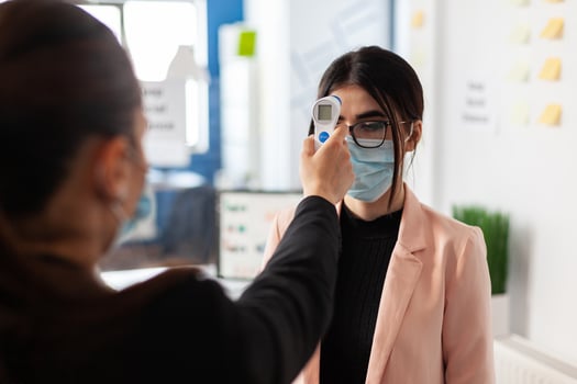 Woman checking colleague temperature using digital thermometer in work office during global pandemic with coronavirus, wearing face mask as safety precaution. New normal virus outbreak. photo
