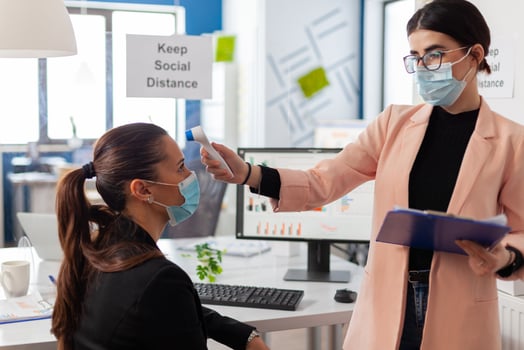 Business woman wearing face mask taking body temperature of colleague in company office using digital thermometer with infrared, during global pandemic with coronavirus, keeping social distancing. photo