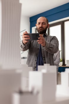 Architect using smartphone to record of architectural maquette of skyscraper to post on social media. Bearded engineer photographing 3d white foam model with smart phone in modern office. photo