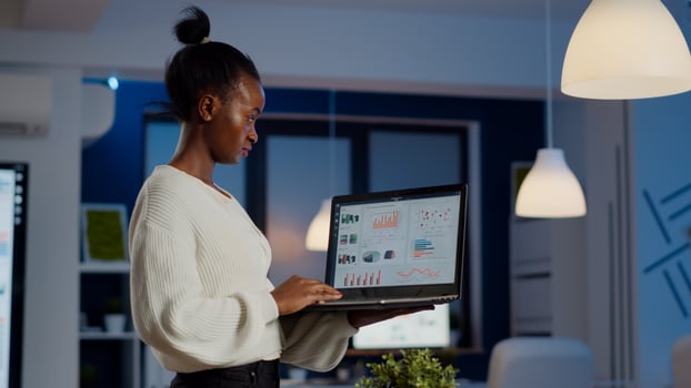 Focused black manager woman checking business report standing in start up office late at night holding laptop. Busy african employee analysing financial statistics overworking writing, searching. photo