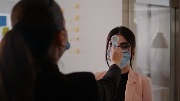 Close up of woman measuring temperature with medical thermometer to prevent covid 19. Coworkers keeping social distance wearing protection face mask at work place during coronavirus global pandemic photo