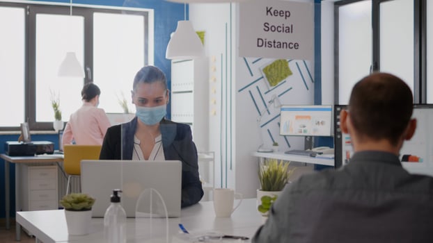 Close up of two coworkers talking about marketing project wearing protective medical face mask sitting in new normal office during coronavirus global pandemic. Team mantain social distancing photo