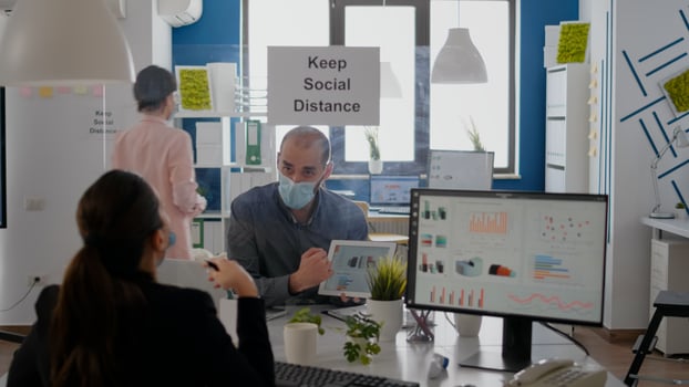 Group of business coworkers with face masks analyzing graphs using digital tablet sitting in new normal workplace. Colleague working in background mantain social distancing photo