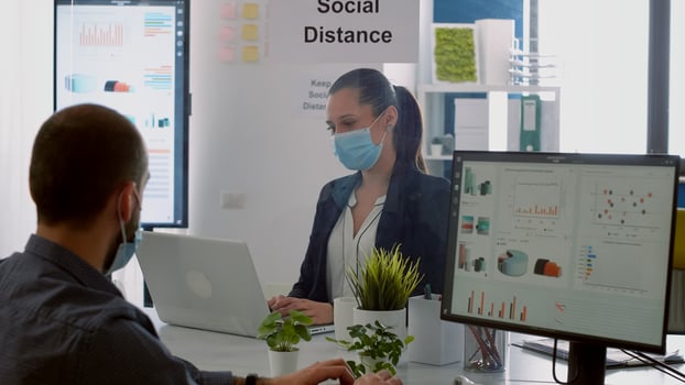 Businessman looking at financial graphs on computer display while talking with coworker sitting in company office. Colleagues with face masks keeping social distancing to prevent covid19 disease photo