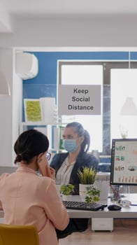 Business woman with medical face mask talking on smartphone with partners while her coworkers working in background respecting social distancing to prevent infection with coronavirus photo