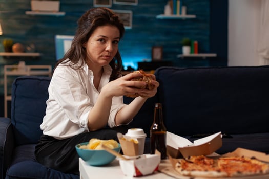 Portrait of relaxed woman looking into camera holding tasty burger in hands during fast food night in living room. Caucasian female enjoying delicious home delivered junk-food. Takeaway delivery photo