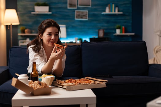Caucasian female holding delicious pizza slice eating takeaway food delivery while watching comedy film on television at night. Woman enjoying junk-food home delivered relaxing on couch photo