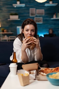 Portrait of caucasian woman looking into camera relaxing on sofa in living room enjoying takeaway food home delivery. Woman eating tasty delicious burger. Fastfood lunch meal order photo