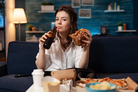 Portrait of woman looking into camera during fastfood lunch meal order relaxing on sofa late at night in living room. Caucasian female enjoying tasty burger, takeaway food home delivered photo