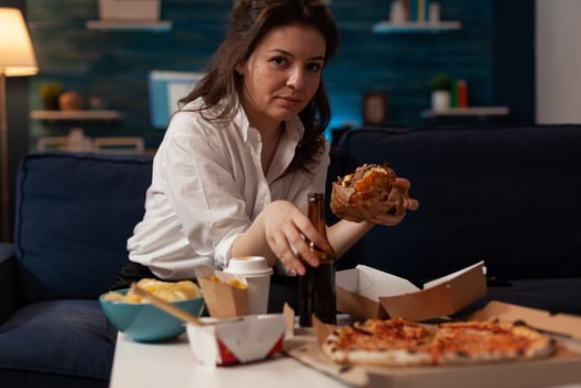 Woman holding beer bottle eating tasty takeaway hamburger while relaxing on couch in front of tv table with fast food menu. Hungry person enjoying junk food sitting on sofa in the evening. photo