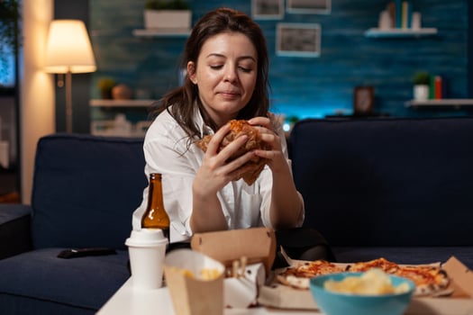 Woman sitting on couch looking at tasty delicious burger delivery dinner in living room in front of television. Person standing at table with takeaway fast food meal large pizza. photo