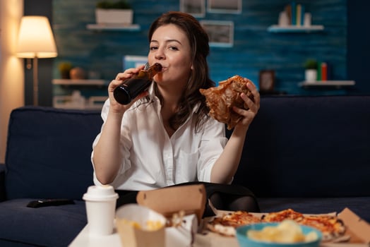 Woman in white shirt drinking beer from bottle while holding a tasty takeaway burger looking at television movie. Person sitting on couch eating delivery hamburger in front of table with fast food. photo