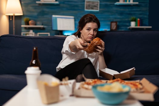 Bored woman sitting on sofa eating tasty delicious burger holding remote switching channels on television in living room. Tired person browsing tv stations while eating takeaway hamburger. photo