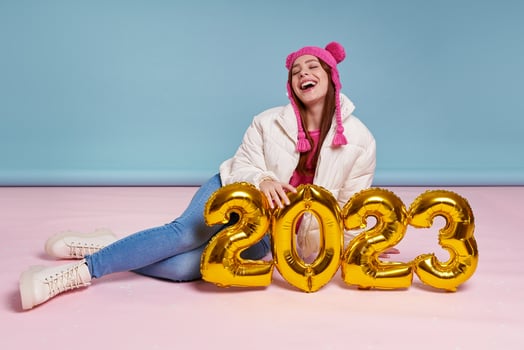 Cheerful young woman in winter coat holding gold colored numbers while sitting against blue background photo