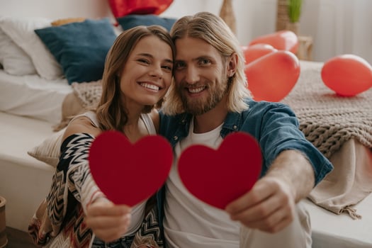 Smiling loving couple embracing and holding heart shape Valentines cards while sitting gin decorated bedroom photo
