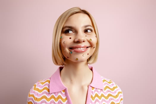 Happy young woman with colorful rhinestones over her face standing against pink background photo