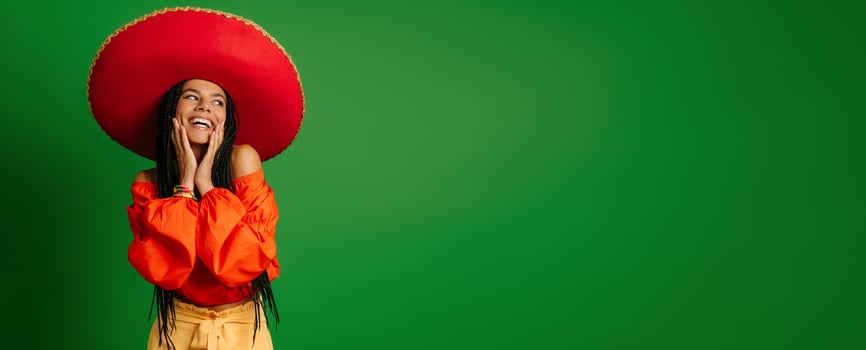 Happy young Mexican woman in Sombrero touching face and smiling against green background photo