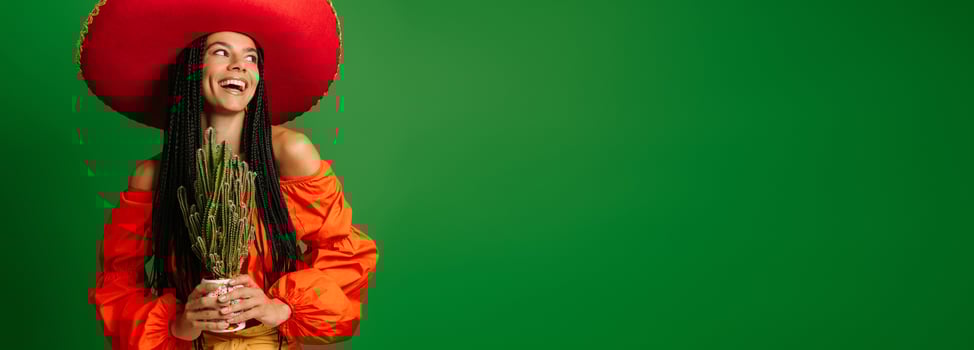 Joyful young Mexican woman in Sombrero holding cactus against green background photo
