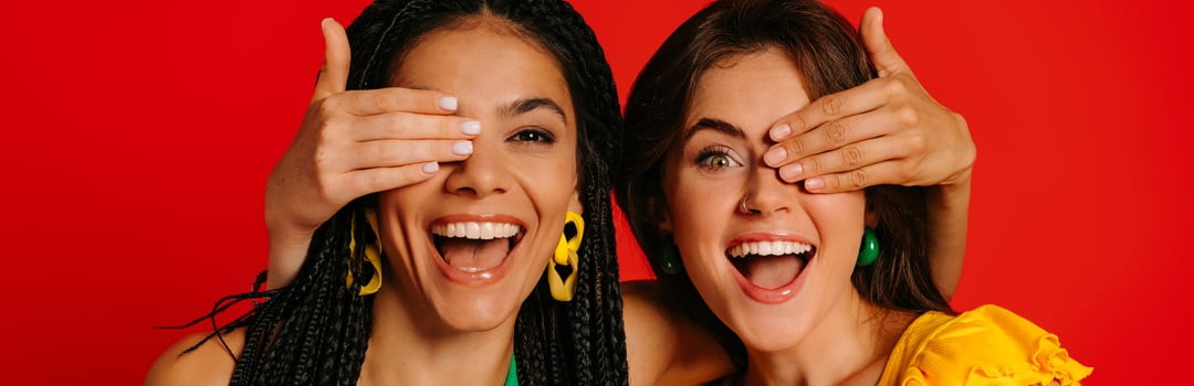 Two happy young women in colorful wear covering eyes to each other and smiling against red background photo